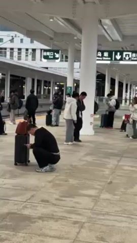 The video shows a train station platform with several people waiting, many with luggage. One person is squatting down near a black suitcase, looking at their phone. Most people are standing and some are looking at their phones. The platform has a light-colored tiled floor and white support columns. There are signs in Chinese above the platform with exit arrows and a location name. A train is visible on the left side of the frame. The scene appears to be indoors, with a roof covering the platform.