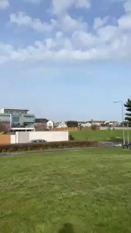 The video depicts a grassy area leading to a row of beach huts under a partly cloudy sky. Initially, the scene shows a green lawn, buildings in the distance, and a sparse scattering of trees. A young person in a pink sweater and dark pants walks across the grass with a light-colored dog, moving towards a row of colorful beach huts on the other side of a parking area. The beach huts are predominantly white with accents of blue, yellow, and gray. Several cars are parked nearby.