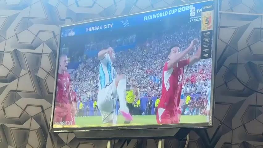 The video displays a television screen showing a soccer match. The screen exhibits the FIFA World Cup 2026 logo and the text KANSAS CITY. On the field, players are in red and white jerseys. One player in a white and light blue jersey is in mid-air, kicking a soccer ball. Another player in a red jersey is positioned to block. The background shows a stadium filled with spectators and several other players on the field. The television set is mounted on a stand, and a wall with a patterned design is visible behind it.