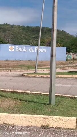 The video shows a roadside view of the Bahia de Plata sign, with a road and landscaping in the foreground. The sign is blue with white lettering and a yellow circle. Behind the sign is a hill covered with vegetation. Trees and a street light are visible on the side of the road.  The sky is blue with some clouds.