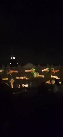 The video shows a night-time projection of animated hands on the facade of a building. The building appears to be a large, classical structure with a dome and several windows. The projected hands are in various poses, some reaching out, others forming shapes. The scene is lit by the projections and some interior lights from the building. Silhouettes of people are visible in the foreground, suggesting an audience watching the projection. The overall atmosphere is one of a public art installation or light show.