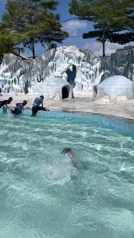 The video shows a swimming pool with three people in or near the water. One person is swimming in the pool. The water is clear and light blue. Two people are near the edge of the pool. One of them is bending over with their hands in the water, and the other is sitting. Behind the pool is a backdrop designed to look like an icy landscape, with structures resembling igloos and artificial ice formations. There are trees on either side of the backdrop. The sky is clear and blue. The scene appears to be outdoors, likely at a water park or recreational facility with a themed area.
