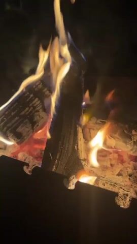 The video shows a close-up view of a campfire. Several logs of wood are burning, with flames and embers visible. The fire is contained within what appears to be a fire pit or grill. The background is dark, suggesting it is nighttime or a dimly lit environment. The flames are flickering and have varying colors, from yellow and orange to hints of blue. The wood is charred and breaking down from the heat. The video is short and focuses solely on the visual aspect of the burning fire.