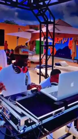 A medium shot reveals a DJ, wearing a tan and camouflage hat, a white t-shirt, and large headphones, standing behind a DJ setup at an outdoor event. The DJ is actively working on a laptop and adjusting equipment, occasionally raising his hands in the air to engage with the music. The setup includes turntables and a laptop, all enclosed in a metal case. Behind the DJ are lounging areas with daybeds, and a colorful mural. There are people in the background, lounging and enjoying the event. Overhead is a black metal trussing system. The lighting is bright and the ambiance suggests a relaxed, upscale outdoor gathering.