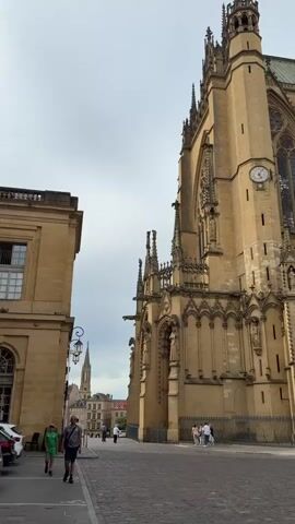 The video shows a streetscape in front of a tall, tan-colored Gothic cathedral with a clock. Two people walk on the cobblestone street, one wearing a green soccer jersey. There are cars parked in front of a building to the left. Other people are walking in the distance. The sky is overcast.