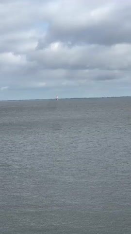 A video captures a wide expanse of choppy gray water under a cloudy sky. In the distance, a small, white lighthouse with red accents is visible on the horizon. The horizon line is flat, suggesting a vast body of water or a very distant coastline. The lighting is subdued, consistent with an overcast day.