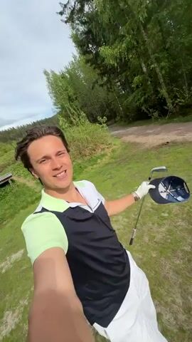 A man takes a selfie on a golf course on a sunny day. He holds up his golf club and cap. The camera pans to show the tee box, and then to a group of three men walking down a path toward the green. The golf course is surrounded by trees and a distant mountain range.
