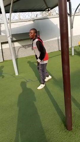 A woman in a red, gray, and black hoodie, gray pants, and white sneakers is attempting to jump rope on a green artificial turf surface. The jump rope is blue. She is smiling and looking towards the camera.  A metal support structure is visible above, casting shadows on the ground.  Another woman wearing gray pants, a patterned tan jacket and white shoes tries to turn the rope for the woman in the hoodie. A third woman in a gray shirt and black pants watches.  A brown post is visible on the right side of the frame.