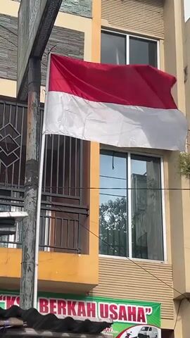 A red and white flag, appearing to be the Indonesian flag, is affixed to a metal pole and is partially obscured by a portion of a building. The building has a beige exterior with windows and a section painted orange. The flag is flapping in the wind. Below the flag, a green banner with red text is visible, reading "BERKAH USAHA". The foreground shows the dark, textured surface of a roof.