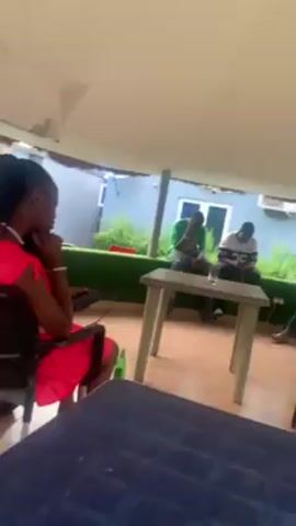 The video shows a group of people sitting in what appears to be an outdoor covered area. One man in a white shirt and blue pants is seated in a chair and makes gestures with his hands. There are tables and chairs scattered around the area. Other people are seated and engaged in various activities like looking at their phones. The outdoor space has a view of a building with windows.