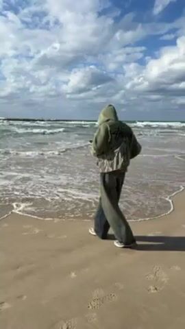 A person walks on a sandy beach towards the ocean. They are wearing a hooded sweatshirt, baggy pants, and sneakers. The sweatshirt is a light olive green color with a graphic print on the back. They are also carrying a woven bag slung across their shoulder. The sky is mostly blue with scattered white clouds. The ocean has white-capped waves, and the water is clear. The person appears to be looking at a phone in their hand as they walk. Footprints are visible in the sand behind them.