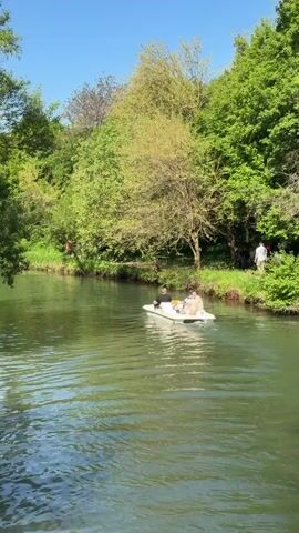 The video shows a calm river with lush green trees lining its banks under a clear blue sky. A white pedal boat with two people on board moves slowly on the water. On the riverbank, a person in red swings from a tree branch over the water, while a small group of people stand nearby, watching. The scene suggests a peaceful day at a park or recreational area.