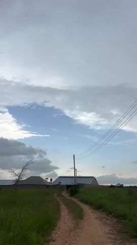 This media file is a vertical video depicting an outdoor scene in Ban, Nigeria. The primary subjects are the natural landscape and the sky. The scene consists of a grassy field in the foreground, with a dirt path winding through it. In the middle ground, there are a few buildings with peaked roofs and brick walls, along with some sparse trees. Power lines are visible in the upper right portion of the frame. The sky is filled with a mixture of clouds, ranging from bright white to dark gray, suggesting a dynamic weather pattern, possibly with an approaching storm.

The video appears to be a passive recording of the environment, with no discernible activity or event taking place beyond the natural movement of clouds and perhaps a gentle breeze rustling the grass. The time of day is not explicitly indicated, but the lighting suggests it could be late afternoon or early evening, given the cloud cover and diffused light. The location cues, such as the style of the buildings and the vegetation, are consistent with a rural or semi-rural setting in Nigeria. There is no visible text in the media file. The overall mood conveyed is one of natural beauty and a sense of anticipation due to the changing weather.