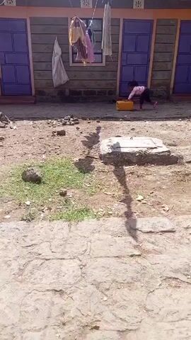 The video shows a small child in a pink shirt and dark pants crawling across a dirt yard. The yard is cluttered with rocks and small patches of grass. In the background is a building with two purple doors and clothes hanging on a line. The child is moving a yellow container.