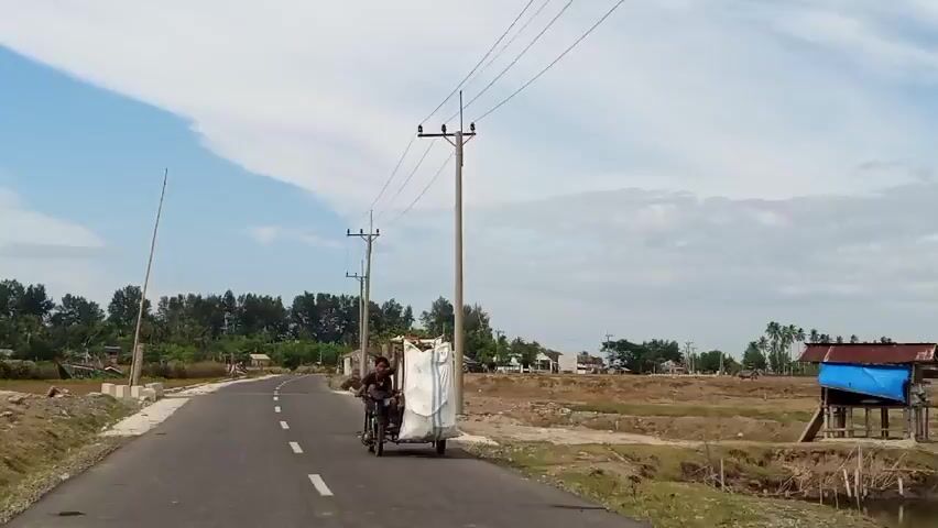 The video shows a paved road with a white dashed line in the center, stretching into the distance. On the right side of the road, a motorcycle with a sidecar is traveling away from the camera. The sidecar is covered with a large white tarp, obscuring its contents. Two people are visible on the motorcycle; one is operating the vehicle, and another is seated behind them. The landscape on either side of the road is mostly dry, with some sparse vegetation and a few scattered buildings. Tall utility poles with power lines run along the right side of the road. In the distance, a line of trees forms a treeline, and the sky is bright blue with some wispy white clouds. The overall impression is that of a rural or semi-rural setting on a sunny day.