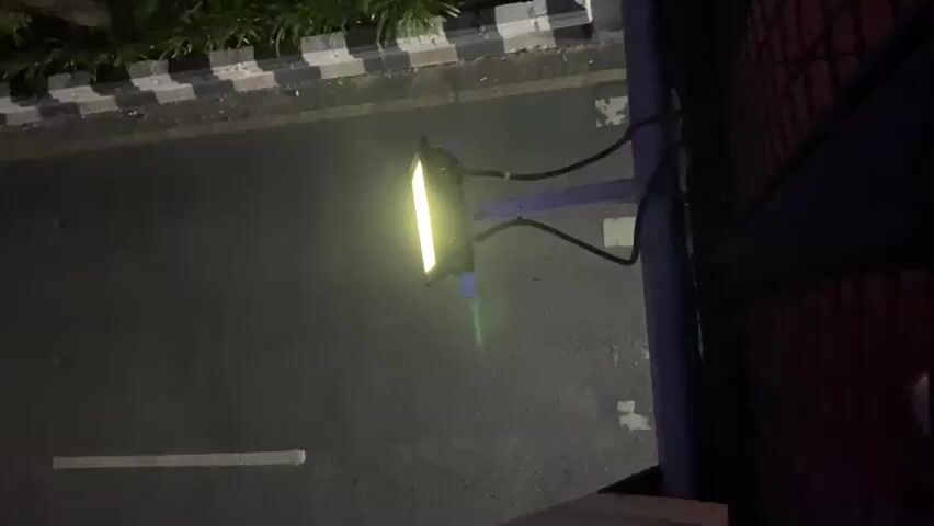 The video is an overhead shot of a street at night. In the foreground, a traffic light pole with a rectangular traffic light hangs over the road. The traffic light is illuminated with a yellow light. A road runs diagonally across the frame, with a striped curb to the left and a brick building to the right.

The first clip shows the traffic light illuminated yellow. In the second clip, a car drives past, its taillights glowing red. In the third clip, a motorcycle rides by, its headlight and taillight visible. The scene appears to be on a city street, given the presence of a traffic light and a building. The time of day is night, as indicated by the darkness and artificial lighting. There is no visible text in the video. The overall mood is calm and ordinary, capturing a typical street scene at night. The location is specified as Banjarbaru, Indonesia.