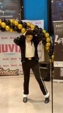 A person dressed as Michael Jackson, wearing a sequined black jacket, white shirt, black pants with white stripes, and white socks with black shoes, performs a dance routine in front of a backdrop featuring balloons and a Muvix Cinemas logo. The person has dark, curly hair and is wearing white gloves. The environment appears to be an indoor event space, possibly a cinema lobby, with a microphone stand and a speaker visible. The lighting is bright, and the overall atmosphere suggests a performance or tribute event.