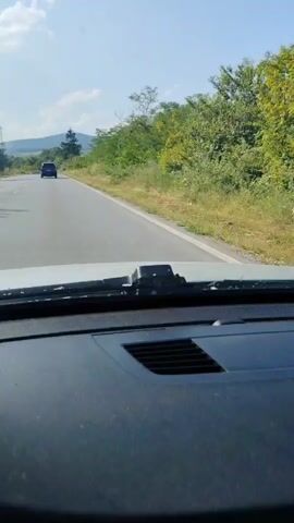 The video, shot from inside a moving vehicle, shows a road trip through a scenic, rural area. The dashboard, windshield, and the base of the rearview mirror are visible in the foreground. A dark-colored car is seen ahead, driving on a two-lane asphalt road.

The road is bordered by lush green vegetation, including trees, bushes, and grass. In the distance, hills or mountains with trees are visible under a clear blue sky with scattered white clouds.

Several road signs are present: a white triangle with a red border and a black arrow indicating a sharp curve to the right, and a circular speed limit sign displaying "70" km/h. A communications tower or cell mast can be seen on the horizon. The weather is sunny and bright. The vehicle is actively driving, following the car in front.