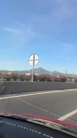 The video shows a car traveling on a highway on a sunny day. The sky is blue with a few clouds. On the side of the highway, there are pink oleander bushes and green trees. In the distance, there are mountains. The car passes a bridge, and several other cars can be seen on the road.