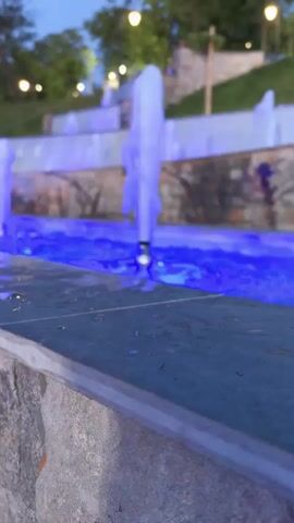 The video shows a series of illuminated fountains in an outdoor setting, likely a park or public space. The water jets are lit with changing colors, transitioning from purple to pink, then orange, and finally green. The background features a grassy slope with trees and stone structures, suggesting a tiered landscape. Several smaller fountains are visible further back, also illuminated. The overall ambiance is tranquil and visually dynamic due to the interplay of light and water. No people are visible in the frame.