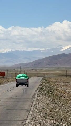 The video features a white pickup truck driving away on a paved road, its bed covered by a prominent bright green tarp. The truck is moving into a vast, mountainous landscape under a bright blue sky dotted with large white, fluffy clouds. Many of the distant mountain peaks are capped with snow.

As the truck recedes, the camera pans slightly to the right, revealing more of the expansive, arid terrain. The roadside is characterized by dry, rocky ground with sparse, light-colored vegetation. In the middle ground, a fence line runs across a wide, flat valley, with what appear to be power lines visible beyond it, leading towards the base of the mountains. The scene conveys a sense of open space and remote travel.

The weather is sunny and clear, suggesting daytime. A red sign with indistinct Chinese characters can be briefly seen on the left side of the road at the beginning of the clip. The overall setting is a rugged, high-altitude environment, likely a rural area within Mainpu township, China.