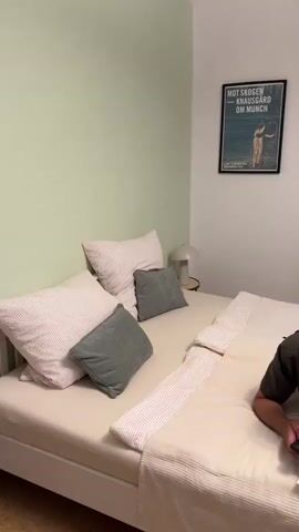 The video shows a bedroom with a man lying on a bed, looking at his phone. The room has a light green wall on the left and a white wall on the right. There is a bed with pillows and blankets. On the wall is a framed picture and a poster. There are two gold palm tree lamps, a red rug, a bookshelf filled with books, and a standing lamp. A chair is visible with clothes and bags on it, and a window with curtains. A robot vacuum cleaner is on the floor. The man is wearing a dark t-shirt and light-colored pants. He is holding his phone and appears to be interacting with it.