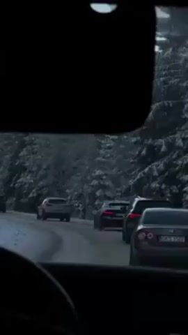 The video shows the view from inside a car driving on a snowy road. The car is behind a line of other vehicles. The trees on either side of the road are covered in snow. The windshield wipers of the car are not moving, and the interior of the car is dark.