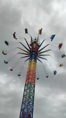 The video shows a tall swing ride with multiple seats hanging from its top, spinning in a circle. The ride is painted in various colors, including blue, green, red, and yellow. People are sitting in some of the swings. The sky behind the ride is overcast with gray clouds.  Some bare tree branches are visible at the bottom of the frame.