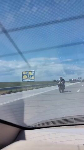 The video shows a view through the windshield of a car traveling on a highway. The sky is blue with some clouds. A billboard is visible in the distance on the right side of the road. A motorcyclist is riding on the right lane of the highway, moving in the same direction as the car. Other vehicles are also visible on the highway in the distance. A tree is visible on the right side of the road.