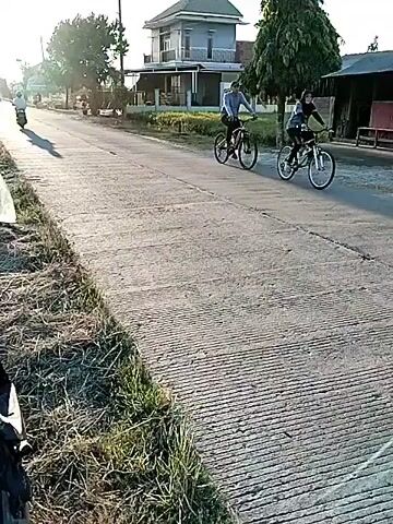 Two individuals are riding bicycles on a paved road. The person in front is wearing a light-colored long-sleeved shirt and dark pants. The person behind is wearing a blue long-sleeved shirt and dark pants. Both are on mountain bikes. The road has a textured surface. To the left of the road, there is grass and a ditch. In the background, there are houses, trees, and utility poles with overhead wires. The sun is visible in the sky, suggesting it is either early morning or late afternoon. The lighting indicates a bright day.