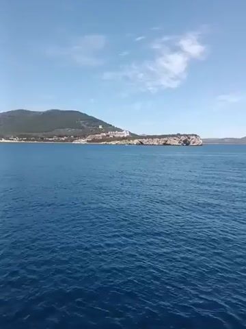 The video shows a wide expanse of calm blue ocean under a clear sky with scattered white clouds. In the distance, a rocky coastline with some buildings can be seen. The camera pans across the water and the coastline. Then, the perspective shifts to show the bow of a boat with a metal gangway. Two people are visible on the boat, one of whom is leaning forward and appears to be speaking or gesturing. The boat is moving through the water, creating gentle ripples. The distant coastline continues to be visible. The lighting suggests it is daytime, likely a sunny day.