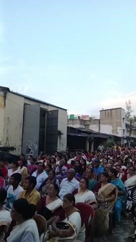 The video captures a large gathering of people, predominantly women, seated outdoors. They appear to be attending an event, possibly a performance or a public address, as they are all facing in the same direction, towards an unseen focal point. The setting seems to be a street or an open area adjacent to buildings. The attendees are dressed in a mix of traditional Indian attire and casual wear. The sky is overcast, suggesting it might be late afternoon or early evening. The atmosphere seems calm and attentive.