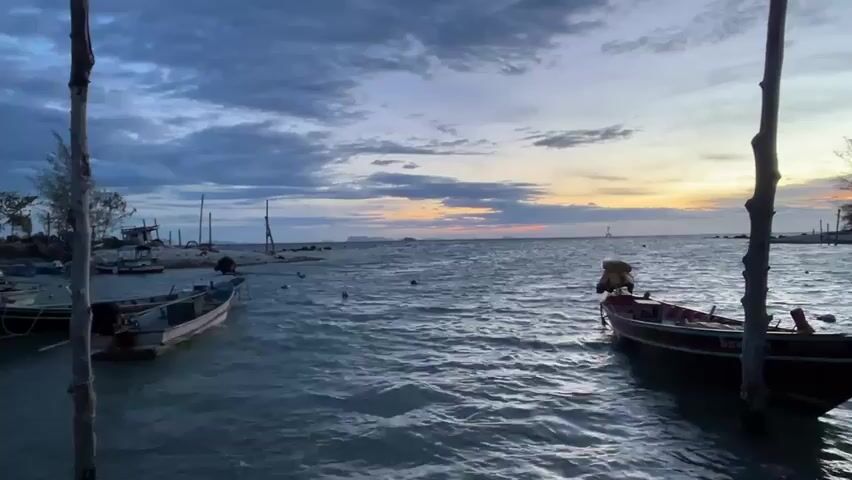 The video shows a view of a coastline with several small fishing boats bobbing in the water. The sky is filled with clouds, with a break in the clouds revealing a hint of sunset on the horizon. Two wooden posts frame the view, adding depth to the scene.