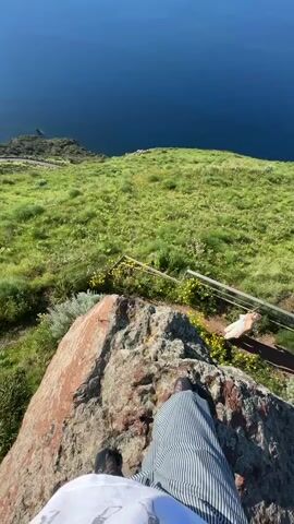 The video opens with a first-person perspective looking down from a rocky cliff edge. The feet of a person wearing striped pants and dark shoes are visible on the reddish-brown rock. Below, a steep grassy slope covered in green foliage and small yellow flowers descends towards the deep blue sea. A wooden railing or fence is visible partway down the slope. Another person in light-colored clothing can be seen lying or sitting on the grass near the fence.

The camera then pans upwards, revealing a breathtaking panoramic view. The vast expanse of the deep blue sea stretches out to the horizon, reflecting the bright sunlight in a dazzling streak. The sky above is a clear, brilliant blue. In the distance, several islands are visible, one large and prominent with a mountainous silhouette, and two smaller ones further away. The foreground remains a verdant hillside, covered in lush green grass and dotted with yellow wildflowers. The sun is high and bright in the sky, indicating daytime. The sound of wind is audible throughout the clip. There is no visible text. The scene depicts a scenic vista from a high point, likely in Lipari, Italy, looking out over the Aeolian Islands.