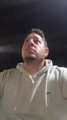 A man is looking at the camera, which is positioned below his face and pointed upwards. He is wearing a light gray hooded sweatshirt with a small green alligator logo on the left chest. He has short dark hair and a mustache, and is outdoors at night. Occasional bright lights pass overhead, presumably from vehicles. There are indistinct buildings and street fixtures in the background. The overall impression is that the man is recording a video of himself while standing on a street.