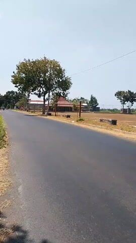 A video captures a view from a moving vehicle on an asphalt road. The road is empty and appears to be in a rural area. On the right side of the road, there is a large, flat, open field that seems to be a sports field or a training ground. There are some low, rectangular structures scattered across the field. Several trees are visible, some in the distance and some closer to the road. There are also two prominent wooden poles with what appear to be lights or decorative elements at the top, marking the edge of the field. A building with a reddish roof and some trees is visible on the left side of the road. The sky is clear and bright, suggesting a sunny day. The video is short and focused on the scenery outside the moving vehicle.