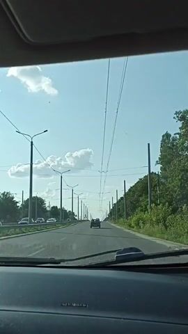 The video shows a view from inside a vehicle looking out through the windshield. The vehicle is on a paved road that appears to be a highway or a major road with multiple lanes. The road stretches into the distance, lined with trees and streetlights on both sides. Overhead, there are numerous wires, likely for public transportation such as trolleybuses. The sky is blue with scattered white clouds, indicating daytime. In the distance, a dark-colored car is driving in the same direction as the camera vehicle. Other vehicles are visible further down the road. The interior of the vehicle is partially visible, including the dashboard and a section of the windshield.