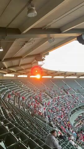 The video shows a panoramic view of a baseball stadium during a game. The camera pans from a close-up of the stadium's roof and a speaker to a wide shot of the field and the stands. The sun is setting, casting a warm glow over the scene, and flares are visible in the sky. Spectators, many wearing red, are seated in the stands. The baseball field is visible, with players on the field and a large scoreboard displaying images. The overall atmosphere appears to be that of a lively sporting event.