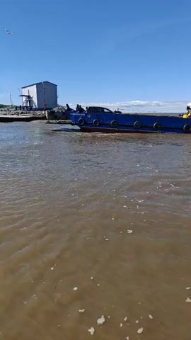 A blue and white boat is traveling through muddy water. The boat is long and narrow, with a white cabin at the back. There are several people standing on the deck of the boat. The sky is blue and clear, and there are some white clouds in the distance. The water is choppy, and there are some whitecaps on the surface. The boat appears to be moving away from the camera.