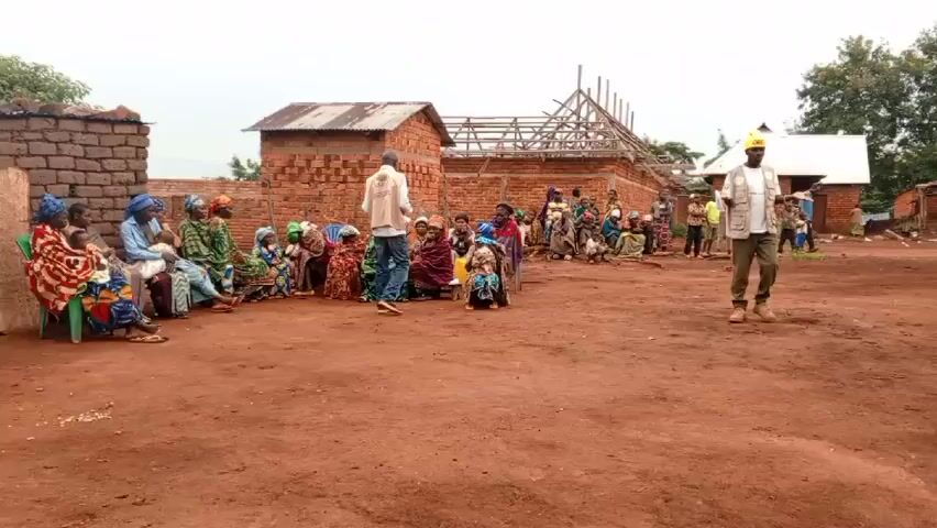 The scene is an open dirt area in a rural village with simple brick buildings with corrugated metal roofs in the background, some of which are under construction. A group of people, mostly women and children, are seated on the ground or small stools, facing a man standing in the center of the space. The man is wearing a white t-shirt, a khaki vest, khaki pants, and a yellow beanie with the word "OBEY" on it. He is gesturing with his hands as if he is addressing the group. Another man in a white shirt and jeans stands near the seated group. A few other people, including some children, stand or wander around in the background. The sky is overcast, suggesting it is daytime. The overall impression is that the man is giving a presentation or instruction to the gathered community members.