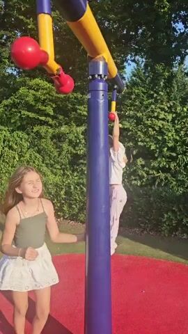 The video shows two young girls at an outdoor playground. The girl in the foreground, facing away from the camera, is looking upwards. She has long, light brown hair and is wearing a green tank top and a white skirt. She is standing on a red circular play surface. In the background, a second girl with blonde hair is hanging from monkey bars attached to a blue and yellow horizontal bar. She is wearing a white t-shirt and light purple pants with a pattern. She is actively swinging from the bars. The playground is surrounded by lush green trees and bushes. The lighting suggests it is daytime, possibly late afternoon, with sunlight casting shadows on the ground.