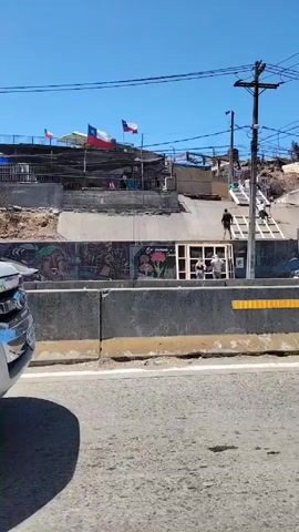The video shows a construction site on the side of a road.  A low concrete barrier separates the road from the site.  A white pickup truck passes by the camera. Three individuals are visible at the site. Two stand near what appears to be a door or window frame, while the third walks along a ramp or stairs constructed on the slope leading up the hillside. Chilean flags are visible above. Overhead, power lines stretch across the blue sky. The video's focus is on the construction activity and the surrounding environment.
