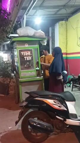 A man wearing an orange t-shirt and a dark baseball cap is standing behind a green and white food cart with the words "TAHL TEK" displayed prominently. He is interacting with a woman who is wearing a dark blue hijab and a maroon dress. The food cart is illuminated by a bright overhead light. To the left of the food cart, a motorcycle is parked, with its front wheel and part of its body visible. The background features a building with colorful striped walls in yellow, green, and red. The scene appears to be outdoors, likely on a street or sidewalk, at night or in dim lighting, given the artificial illumination.