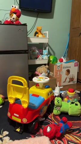 The video shows a cluttered playroom with various toys. In the foreground, a red and yellow ride-on car is parked on a colorful rug. To the right of the car, a stuffed Spiderman toy lies on its back. Further back, a green stuffed Creeper toy from Minecraft is visible, along with a small toy airplane. A tiered shelf unit stands against the wall, filled with stuffed animals, including Elmo on top of a refrigerator to the left. A box of diapers is also visible near the shelf. A television screen is mounted on the wall above the shelf. The room appears to be a child's play area, with toys scattered on the floor and shelves.