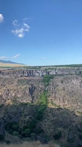 The media file is a landscape video showcasing a canyon with a waterfall in Ohanavan, Armenia. The scene is a natural, rugged environment dominated by steep, rocky cliffs and lush green vegetation. The waterfall is a prominent feature, cascading down the cliff face into the canyon below. The top of the canyon is a plateau with sparse trees and a clear blue sky with a few wispy clouds. The time of day appears to be daytime, likely late morning or early afternoon, given the bright sunlight casting shadows on the canyon walls. The weather seems pleasant and sunny. There are no visible people or man-made objects, emphasizing the natural beauty of the location. The overall mood is serene and majestic, highlighting the grandeur of the natural landscape. There is no visible text within the media file.