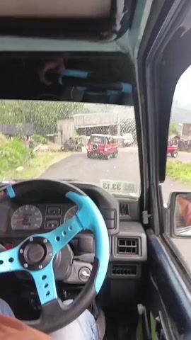 The video is taken from inside a vehicle looking out the passenger side window and through the windshield. The driver's hands are visible on the steering wheel, which has a light blue rim. The dashboard of the vehicle is also visible. Outside the vehicle, it appears to be a rural or suburban area with buildings, trees, and piles of tires. There are other vehicles visible on the road, including a red SUV in the distance. The sky is overcast, and there appears to be rain or spray on the windshield. The overall impression is that the vehicle is driving on a road in a somewhat unkempt environment.