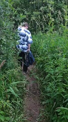 A person wearing a short-sleeved shirt with a pattern of hippopotamuses and dark pants is walking down a dirt path through tall green grass and foliage. They are holding a dark blue item in their left hand. The path appears narrow, with vegetation crowding its sides. The scene is outdoors, with trees visible in the background. The sky is overcast. The camera pans slightly to the right, showing more of the dense vegetation and the sky.