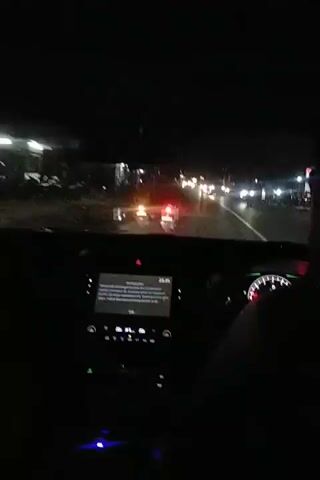 The video shows a view from inside a car driving at night. The dashboard is visible, with a screen displaying a message and various controls illuminated. Outside, there are other vehicles on the road, including motorcycles with their taillights visible. Streetlights and other lights illuminate the roadside, suggesting a populated area. The overall environment is dark, with the car's headlights illuminating the road ahead.