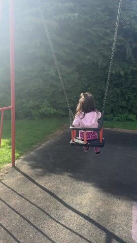 The video shows a young girl sitting on a swing in a park. She is wearing a light pink hooded sweatshirt and patterned pink and purple leggings. The swing has a red frame and a black seat, and hangs from metal chains attached to a red metal frame. The girl is swinging back and forth, seen from the back. Behind the swing is a lush green hedge. The ground beneath the swing is black rubber surfacing with the shadows of the swing frame cast upon it. The camera then pans to a grassy area with playground equipment in the distance. The day appears sunny.