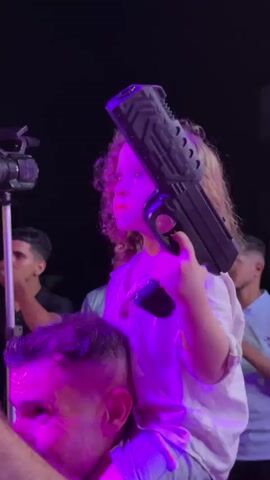 A child sits on an adult's shoulders, holding a large black toy gun. The child has curly blonde hair, wears a white shirt, and has red lipstick on. The scene is dimly lit, with colorful stage lights casting green and blue hues. In the background, other people are visible, some clapping. The environment appears to be an outdoor event or performance space, with a stage and decorations overhead. The camera pans to show a crowd of people dancing and enjoying themselves under the lights.